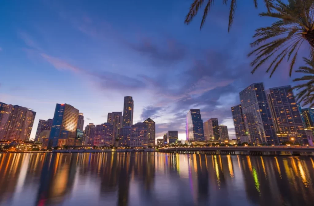 Skyline de Miami ao entardecer refletido na água, com prédios modernos e iluminação urbana